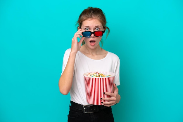 Joven inglesa aislada de fondo azul sorprendida con gafas 3d y sosteniendo un gran cubo de palomitas de maíz