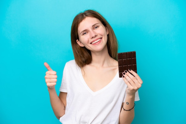 Joven inglesa aislada de fondo azul tomando una tableta de chocolate y con el pulgar hacia arriba