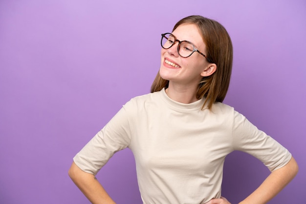 Joven inglesa aislada de fondo morado posando con los brazos en la cadera y sonriendo