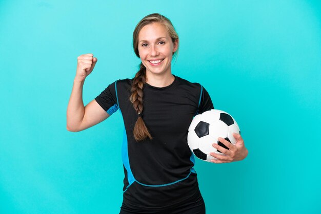 Joven inglesa aislada sobre fondo azul con balón de fútbol celebrando una victoria