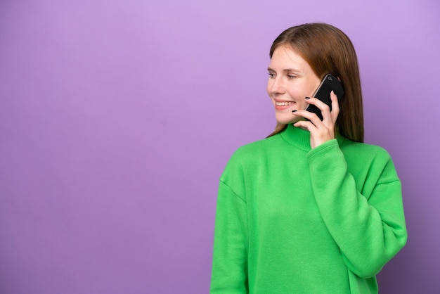 Joven inglesa aislada sobre fondo púrpura manteniendo una conversación con el teléfono móvil con alguien