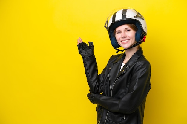 Joven inglesa con un casco de motocicleta aislado en un fondo amarillo extendiendo las manos a un lado para invitar a venir