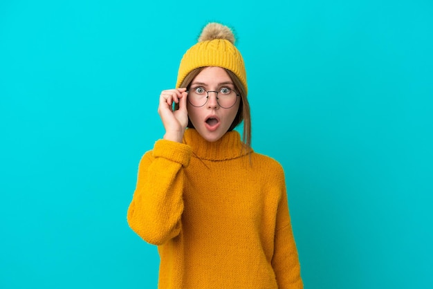 Joven inglesa con chaqueta de invierno aislada de fondo azul con gafas y sorprendida