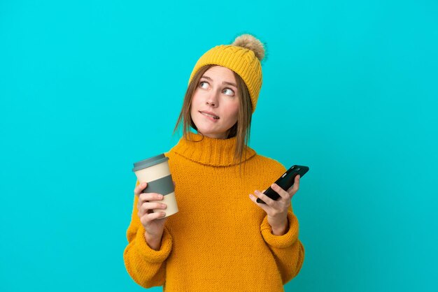 Joven inglesa con chaqueta de invierno aislada de fondo azul sosteniendo café para llevar y un móvil mientras piensa en algo