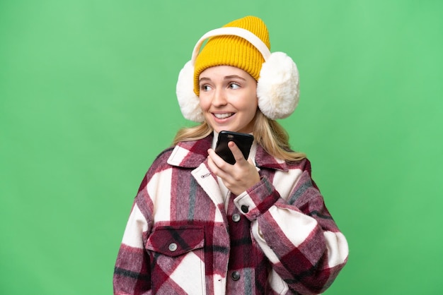 Joven inglesa con manguitos de invierno sobre un fondo aislado manteniendo una conversación con alguien con el teléfono móvil