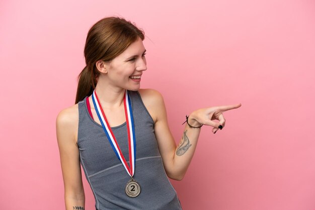 Joven inglesa con medallas aisladas de fondo rosa señalando con el dedo al lado y presentando un producto