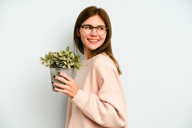 Una joven inglesa que sostiene una planta aislada de fondo azul mira a un lado sonriente, alegre y agradable.
