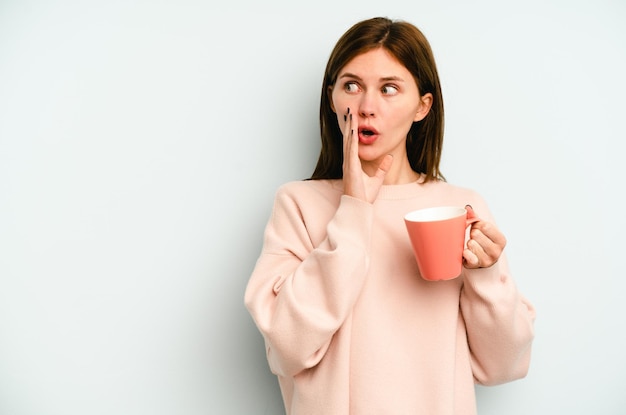 Una joven inglesa que sostiene una taza aislada de fondo azul dice una noticia secreta de frenado en caliente y mira a un lado