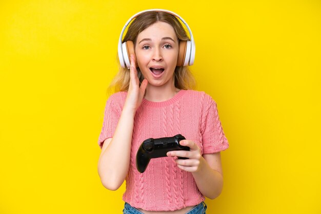 Una joven inglesa rubia jugando con un controlador de videojuegos aislado en un fondo amarillo con una expresión facial sorprendida y sorprendida