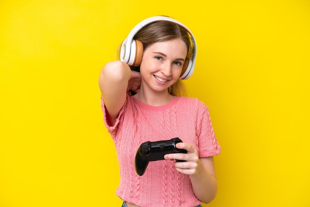 Una joven inglesa rubia jugando con un controlador de videojuegos aislado en un fondo amarillo riéndose