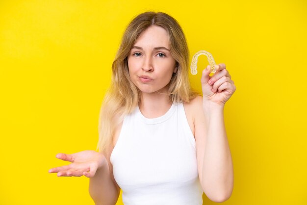 Una joven inglesa rubia sosteniendo aparatos invisibles aislados en un fondo amarillo haciendo gestos de duda mientras levanta los hombros