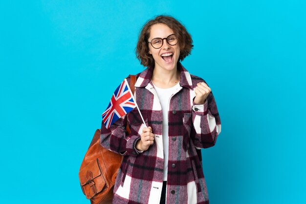 Joven inglesa sosteniendo una bandera del Reino Unido aislada en la pared azul celebrando una victoria en la posición ganadora