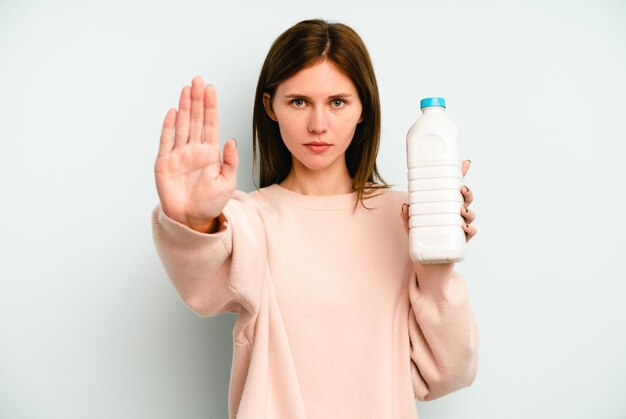 Joven inglesa sosteniendo una botella de leche aislada de fondo azul de pie con la mano extendida mostrando la señal de alto, impidiéndote.