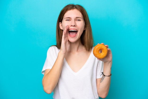 Joven inglesa sosteniendo un donut sobre un fondo azul aislado gritando con la boca abierta