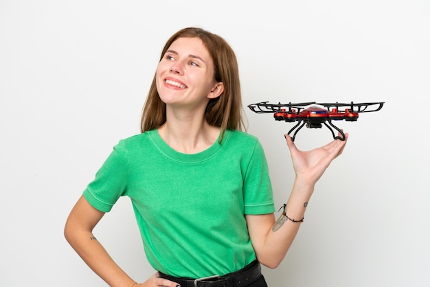 Joven inglesa sosteniendo un dron aislado de fondo blanco posando con los brazos en la cadera y sonriendo