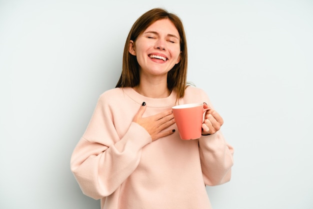 Joven inglesa sosteniendo una taza aislada de fondo azul se ríe a carcajadas manteniendo la mano en el pecho