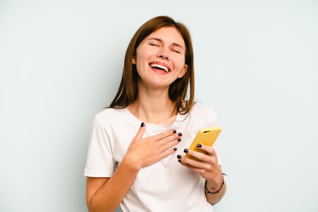 Joven inglesa sosteniendo un teléfono móvil aislado de fondo azul se ríe a carcajadas manteniendo la mano en el pecho