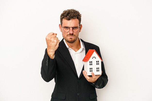 Joven inmobiliaria caucásica sosteniendo una casa de juguete aislada sobre fondo blanco mostrando el puño a la cámara, expresión facial agresiva.