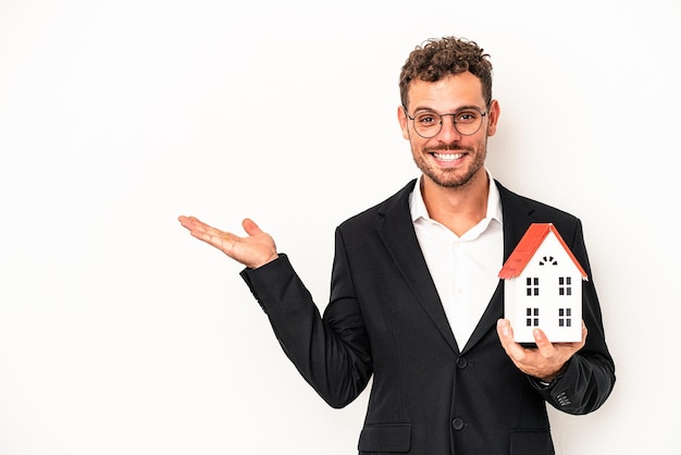 Joven inmobiliaria caucásica sosteniendo una casa de juguete aislada sobre fondo blanco que muestra un espacio de copia en una palma y sosteniendo otra mano en la cintura.