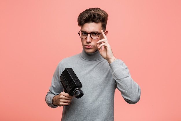 Joven intelectual sosteniendo una cámara de cine apuntando su sien con el dedo, pensando, centrado en una tarea.