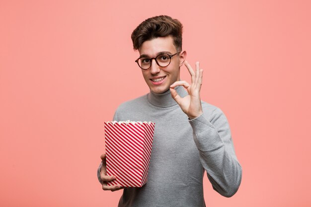 Joven intelectual sosteniendo un cubo de palomitas de maíz sorprendido apuntando a sí mismo, sonriendo ampliamente.