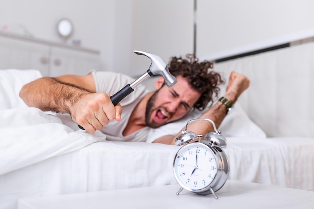 Joven intenta romper el despertador con un martillo, Destroy the Clock. Hombre acostado en la cama apagando un despertador con un martillo por la mañana a las 7 de la mañana.