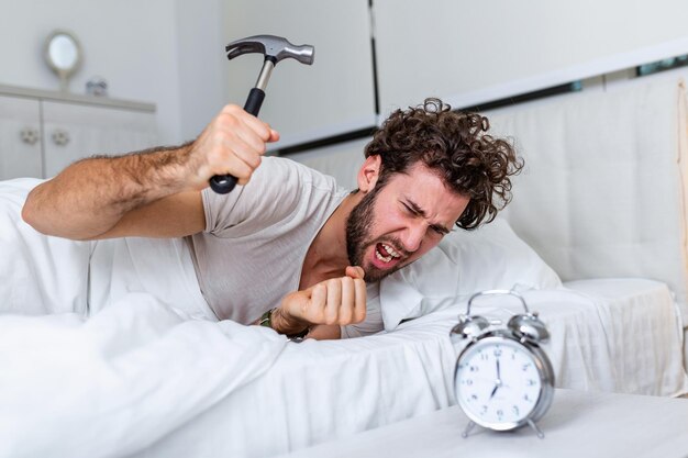 Joven intenta romper el despertador con un martillo, Destroy the Clock. Hombre acostado en la cama apagando un despertador con un martillo por la mañana a las 7 de la mañana.
