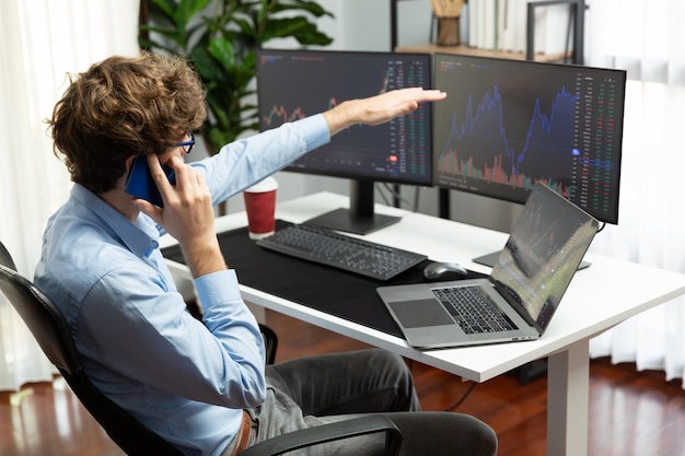 Un joven inversor de acciones exitoso llamando por teléfono inteligente a un socio de negocios con una pantalla de portátil de inversión dinámica en el gráfico actual del mercado en tiempo real en la oficina moderna de Gusher.