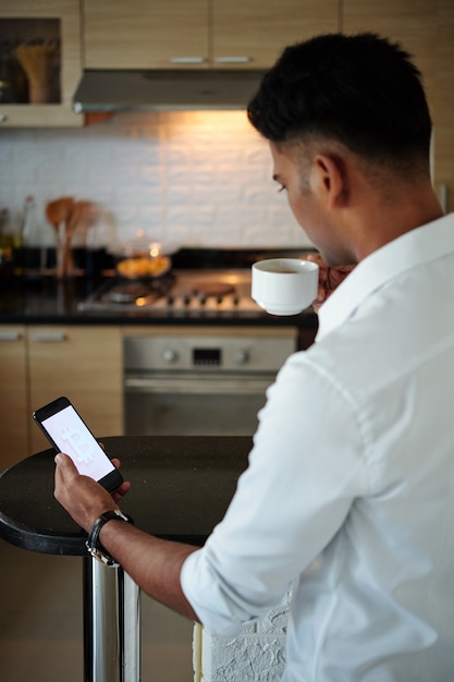 Foto joven inversor bebiendo una taza de café por la mañana en la cocina y comprobando el precio de bitcoin y las predicciones a través de la aplicación en el teléfono inteligente