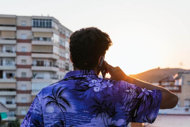 Un joven irreconocible boca arriba en el centro de la imagen con una camisa azul hawaiana hablando por teléfono con su teléfono inteligente durante la puesta de sol Concepto de teletrabajo criptomoneda