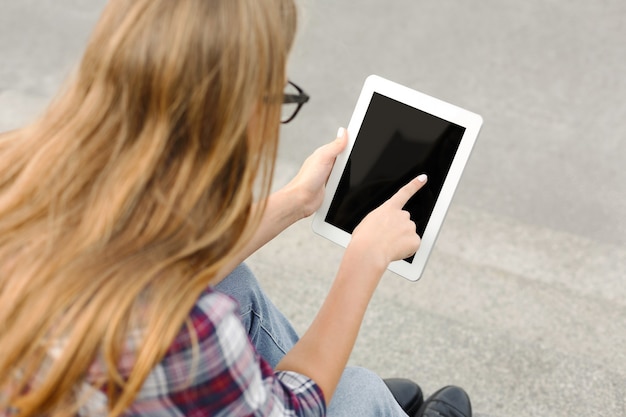 Joven irreconocible sentada al aire libre en las escaleras de la universidad con tableta digital, escribiendo, navegando por internet, preparándose para los exámenes, vista posterior. Tecnología, educación y concepto, espacio de copia.