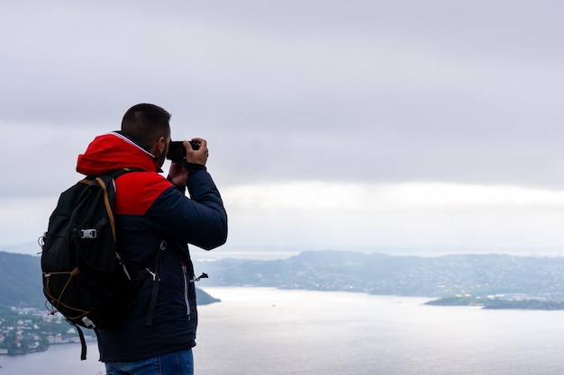 Joven irreconocible con su mochila en la espalda y fotografiando la ciudad de Bergen y el fiordo en Noruega