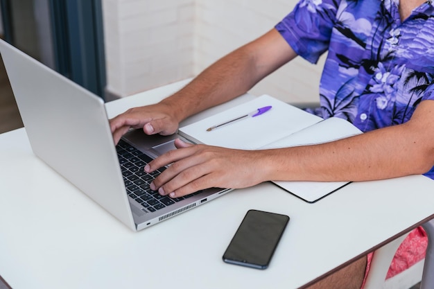 Joven irreconocible trabajando con una computadora portátil en la terraza en traje de baño y camisa hawaiana azul sobre fondo de ladrillo blanco concepto criptomoneda de teletrabajo