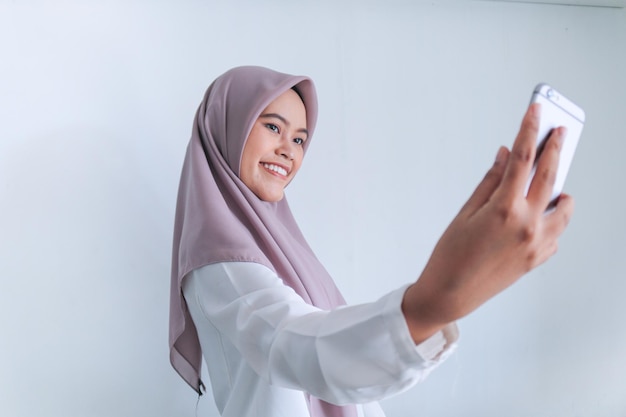 Una joven islámica asiática con pañuelo en la cabeza sonríe y se toma selfies con su teléfono móvil Mujer indonesia de fondo gris