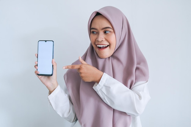 Una joven islámica asiática que lleva un pañuelo en la cabeza señala con el dedo en la pantalla blanca del teléfono con una sonrisa y un sentimiento feliz Mujer indonesia de fondo gris