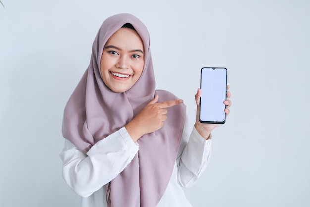 Una joven islámica asiática que lleva un pañuelo en la cabeza señala con el dedo en la pantalla blanca del teléfono con una sonrisa y un sentimiento feliz Mujer indonesia de fondo gris