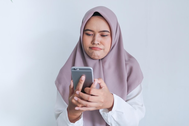 La joven islámica asiática que lleva velo está triste y llora por lo que ve en el teléfono inteligente Mujer indonesia de fondo gris