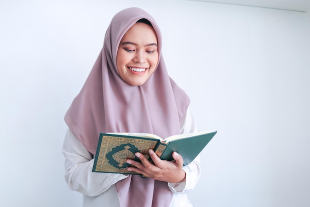 Una joven islámica asiática con velo reza o lee el Corán, el libro sagrado del Islam, con una sonrisa y una cara seria Mujer indonesia de fondo gris