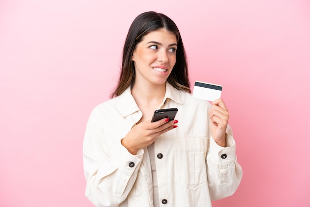 Joven italiana aislada de fondo rosa comprando con el móvil con tarjeta de crédito mientras piensa