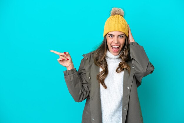 Joven italiana con chaqueta de invierno y sombrero aislado de fondo azul sorprendida y señalando con el dedo al costado