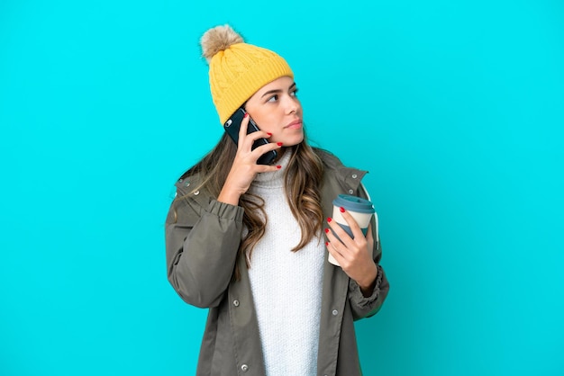 Joven italiana con chaqueta de invierno y sombrero aislado de fondo azul sosteniendo café para llevar y un móvil