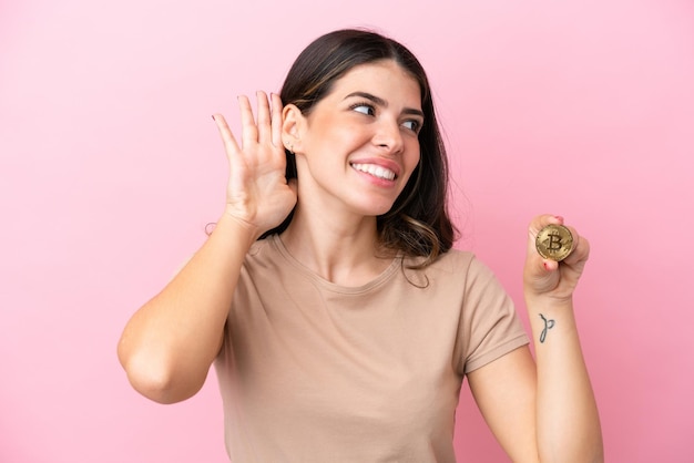 Joven italiana sosteniendo un Bitcoin aislado de fondo rosa escuchando algo poniendo la mano en la oreja