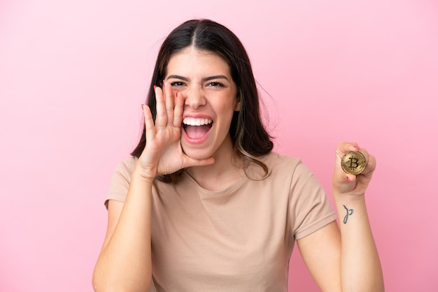 Joven italiana sosteniendo un Bitcoin aislado de fondo rosa gritando con la boca abierta