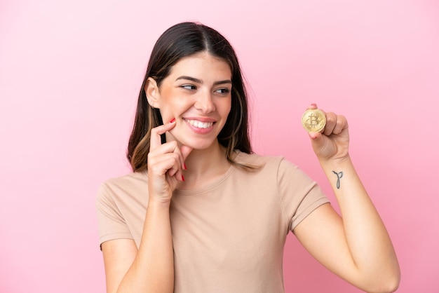 Joven italiana sosteniendo un Bitcoin aislado de fondo rosa pensando en una idea mientras mira hacia arriba