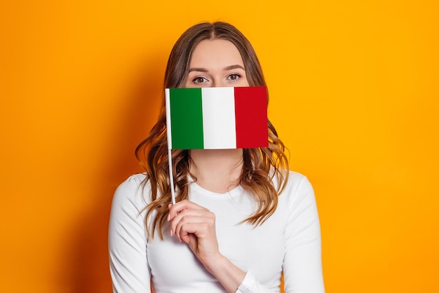 Una joven italiana vestida de blanco se cubre la cara con una pequeña bandera italiana aislada de fondo naranja