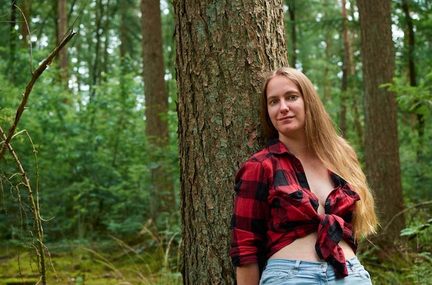 Una joven italiana vistiendo una franela roja con jeans y apoyado en un árbol en un bosque