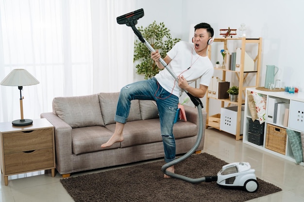 Joven japonés asiático divirtiéndose durante la limpieza de la casa. un tipo guapo se relaja jugando con una aspiradora en la sala de estar mientras usa auriculares y baila con música. cuerpo móvil doméstico masculino