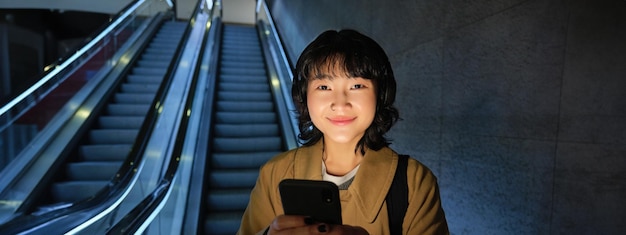 Joven japonesa de pie con un teléfono inteligente bajando una escalera mecánica con un teléfono móvil viajando