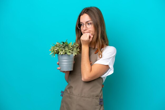 Joven jardinera caucásica sosteniendo una planta aislada de fondo azul con dudas