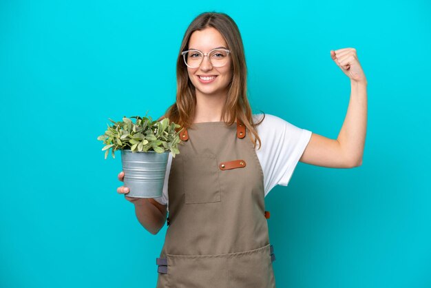Joven jardinera caucásica sosteniendo una planta aislada de fondo azul haciendo un gesto fuerte